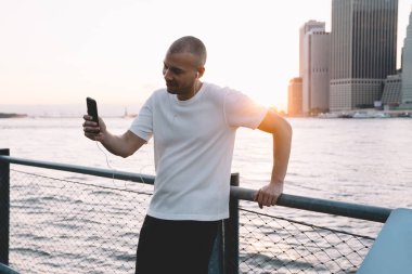 Genç, yakışıklı, kulaklıklı, akıllı telefonuyla selfie çeken ve gün ışığında nehir ve şehrin zincire bağlı metal çitlere yaslanan yakışıklı, etnik bir adam.