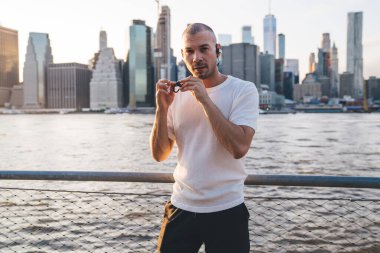 Kısa saçlı spor elbiseli genç adam demir parmaklıkların yanında duruyor gökdelenlerin arka planında çit örüyor, kablosuz kulaklık takıyor ve kameraya bakıyor.