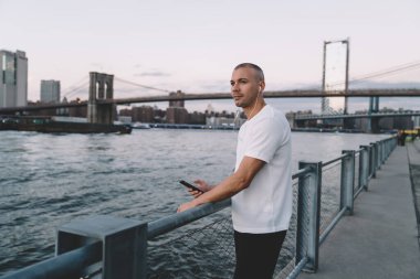 Kısa saçlı, spor elbiseli, akıllı telefon ve kulaklıklı, nehir kenarında zincirli metal çitlere dayanan yakışıklı adamın yan görüntüsü.