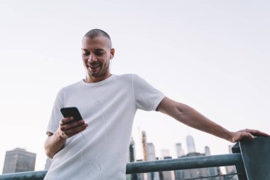 Aşağıdan gülümseyen, beyaz tişörtlü, kulaklıkla müzik dinleyen ve metal çitlere yaslanarak akıllı telefon kullanan genç bir adam. 