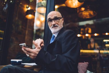 Gözlüklü ve takım elbiseli, akıllı telefonlu yaşlı yakışıklı bir erkek cama yaslanmış ve caddedeki kafede kameraya bakıyor.