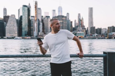 Kısa saçlı, kısa saçlı, kulaklıklı ve akıllı telefonlu yakışıklı genç adam nehir kenarında çitlere yaslanırken gözlerini kaçırıyor.