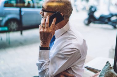 Beyaz tişörtlü, gri kısa saçlı, akıllı telefondan şehrin arka planında oturan ve konuşan yaşlı adamın yan görüntüsü.