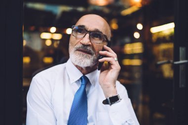 Gözlüklü, takım elbiseli, beyaz gömlekli, akıllı telefondan konuşan, camlara bakan yaşlı, kısa saçlı, yakışıklı bir erkek.
