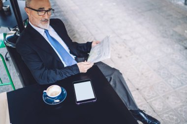 Yukarıdan bakıldığında kendine güvenen üst düzey yöneticinin masada boş ekranlı akıllı telefonuyla oturup gazete okurken kameraya baktığı görülüyor.