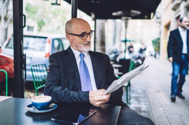 Ofis kıyafeti giyen, kravat takan ve gözlüklü neşeli, son sınıf kel bir adam sokak kafesinde oturmuş, bulanık şehir geçmişi hakkında gazete okuyor. 