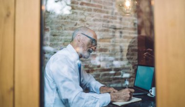 Kafede dizüstü bilgisayarını incelerken, not defterine not alan gözlüklü, odaklanmış yaşlı bir adamın yan görüntüsü.