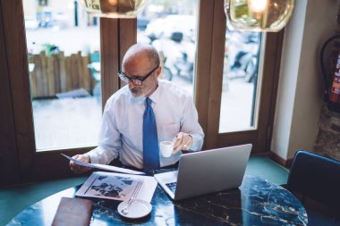 Yukarıdan bakıldığında, gözlüklü, elinde bir fincan kahve kağıdıyla masada, pencerenin yanında dizüstü bilgisayarlı bir şekilde otururken görülüyor. 