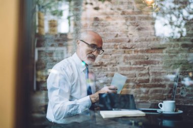 Gözlüklü, yaşlı, kel bir adamın resmi kıyafetlerini incele. Kafede dizüstü bilgisayarla otururken not al.