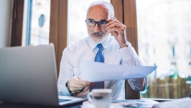 Üst düzey yönetici gözlüğü düzeltiyor ve bulanık arka planda pencerenin yanında kahve fincanı ile masada otururken laptop ekranına bakıyor.