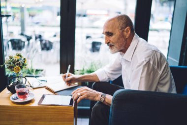 Beyaz gömlekli ve not defterine sakallı kıdemli işadamının yan görüntüsü. Tableti karıştırırken ve arka plan penceresindeki kafede oturmuş çay içerken. 