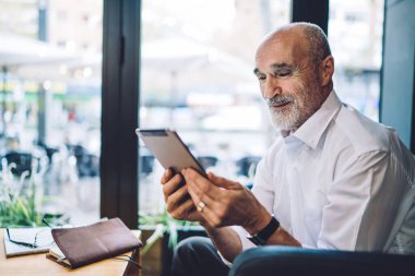 Gülümseyen yaşlı kel adam resmi kıyafetler içinde modern tablet kullanarak kafedeki koltukta zevkle oturuyor.