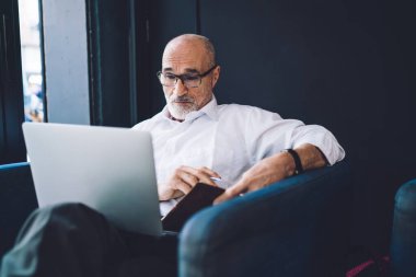 Yaşlı, sakallı, beyaz gömlekli, siyah pantolonlu, dizüstü bilgisayarlı, pencerenin yanında oturan yaşlı adam.