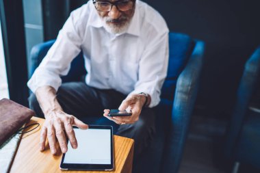 Resmi kıyafetler ve gözlüklü yaşlı adam rahat koltukta oturuyor, akıllı telefona bakıyor ve boş tablet ekranına dokunuyor. 