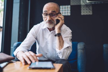 Kel, gri sakallı, gözlüklü, beyaz gömlekli, mavi sandalyede oturan ve telefonla konuşurken tablet üzerinde gezinen yaşlı bir erkek.
