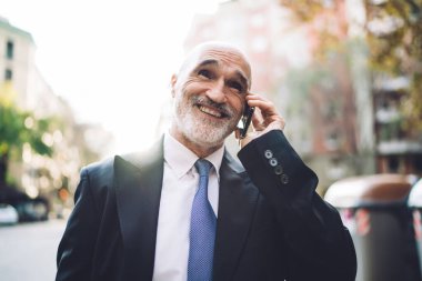 Resmi kıyafetli, yakışıklı, yaşlı ve beyaz sakallı bir erkek sokakta yürürken telefonla konuşuyor. Bulanık binaların arka planına bakıyor. 