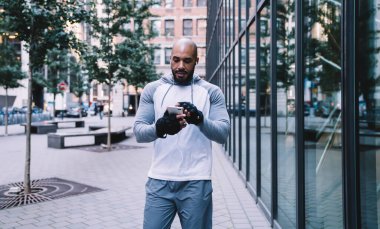 Atletik kel sakallı Afro-Amerikan sporcu kulaklıkla müzik dinliyor ve modern ofis binasının yanındaki kaldırımda dikilirken akıllı telefonuna bakıyor.