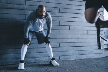 Gri spor giyimli, kel ve yorgun bir adam duvara yaslanmış, dizlerinin üstüne çökmüş ve antrenman sırasında fotoğraf makinesiyle ekin fotoğrafçısına bakıyor.