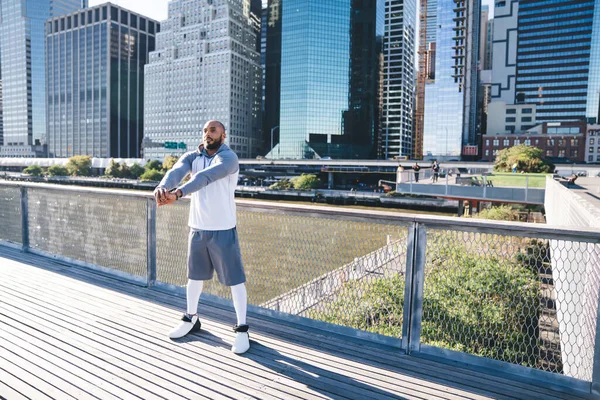 身穿运动服的秃头非洲裔美国人 穿着运动服 伸展着胳膊 然后站在桥上跑步 背景是现代市中心 — 图库照片