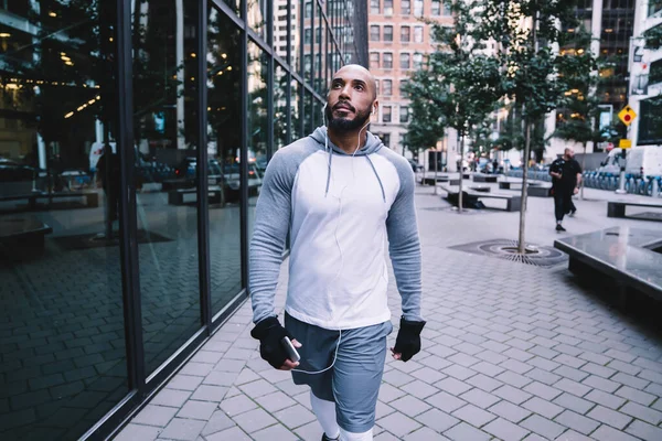 Athletic bald man full of confidence in sportswear walking at street in earphones with smartphone in hand and looking away