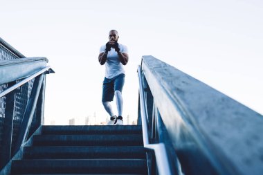 Tişörtlü ve şortlu kel sakallı Afrikalı Amerikalı savaşçı bulutsuz gökyüzüne karşı gölgeli boks yapıyor.