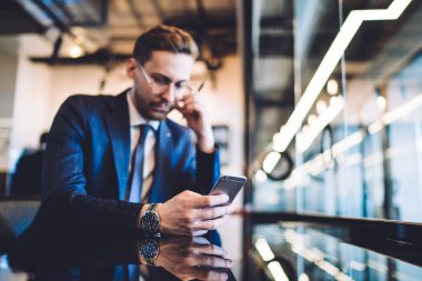 Kol saati takan, bir eliyle gözlüğe dokunan ve diğer eliyle modern tasarım işyerinde akıllı telefondan yazan damalı resmi takım elbiseli bir işadamının yumuşak odağı.