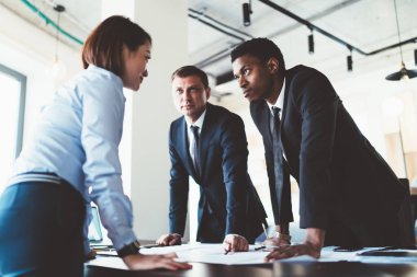 Çok ırklı genç iş arkadaşları ellerinde cihazlar ve belgelerle masanın yanında duruyor ve proje planını şirket ofisindeki toplantı odasında analiz ediyorlar.