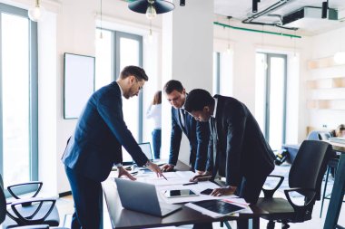 Takım elbiseli çeşitli iş arkadaşlarının yan görüntüsü modern ofis odasında masa başında dururken iş projesi fikirlerini paylaşıp tartışıyorlar.