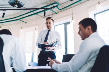 Gözlüklü ve not defteriyle şık kıyafetler giyen dalgın bir çalışanın ayakta durup masasında bulanık, suratsız bir iş arkadaşı üzerinde çalışırken stratejik planlar geliştirmesinden.