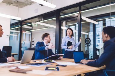 Çok yönlü başarılı insanlardan oluşan bir grup büyük açık ofiste klasik giyinmiş bir adam kapıdaki iş kadınının oturmasını öneriyor.