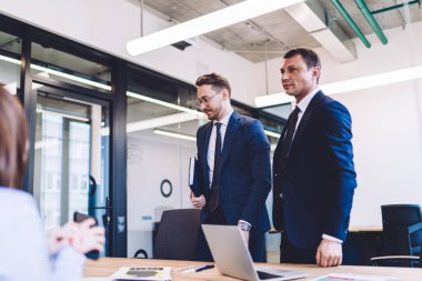 Ciddi, kendine güvenen yönetici ve not defteri olan erkek meslektaşı iş arkadaşlarıyla iş görüşmesi için masada birlikte duruyorlar.