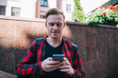 Kızıl sakallı genç erkek işaretli kırmızı ve siyah gömlekli. Yazın şehirde gezintiye çıkan cep telefonuyla sörf yapıyor. 