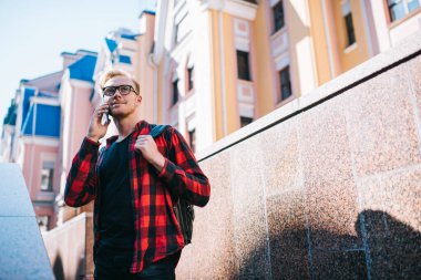 Entellektüel gülümseyen kızıl saçlı erkek işaretli kırmızı ve siyah gömlekli cep telefonuyla konuşuyor şehirdeki granit barajın üzerinde duruyor.