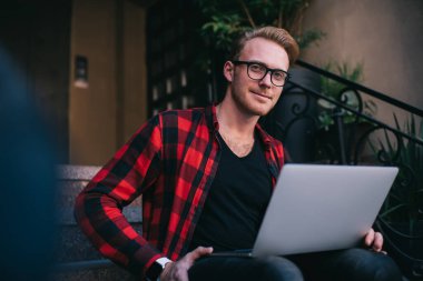 Günlük kıyafetleri ve gözlükleri olan yakışıklı bir adam dizüstü bilgisayar kullanıyor. Yeni başlayanlar için evde oturup kameraya bakıyor.