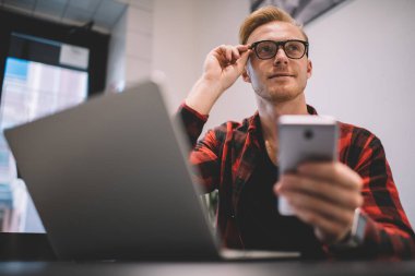 Gündelik genç erkek dizüstü bilgisayar kullanıyor. Yeni başlayan telefonlar üzerinde çalışıyor. Gözlükleri ayarlıyor.