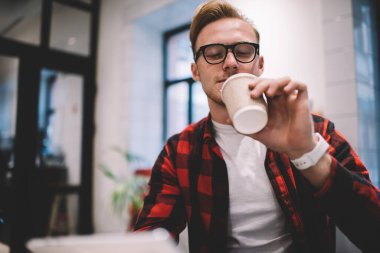 Rahat giyinen ve gözlüklü adam işten dinleniyor. Kahve barında oturmuş sıcak içeceğin tadını çıkarıyor.