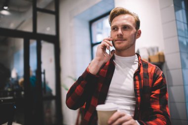 Rahat giyinen genç bir erkek, arkadaşlarıyla konuşuyor. Akıllı telefon kullanıyor. Bir fincan sıcak içecek içiyor, kafeteryada dinleniyor.
