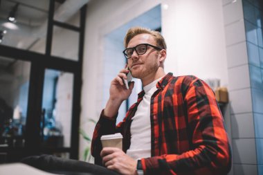 İş yerinde kahve içip kameraya bakarken kareli tişörtlü neşeli adamın cep telefonundan araması.