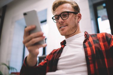 Aşağıdan, gözlüklü barışçıl bir genç adam modern akıllı telefonu dikkatle kullanıyor.