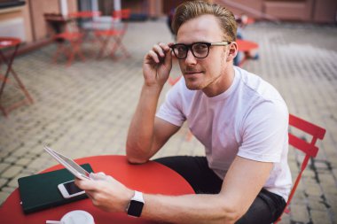 Kendine güveni olan, akıllı saat takan, tablet kullanan, sokak kafesindeki kameraya bakan, kendine güvenen genç bir adam.