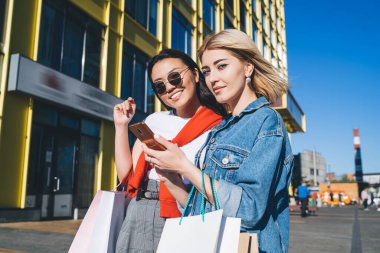 Güneş gözlüklü zeki bayan alışveriş merkezinin önünde alışveriş torbaları taşıyan genç sarışınla takılıyor ve kameraya bakıyor.