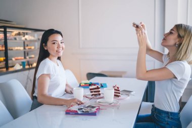 Beyaz tişörtlü çok ırklı kadınlar kahve fincanlarında kek fincanları, kahve fişleri ve arka planda tezgah ile masada oturuyorlar.