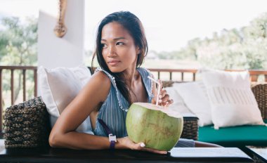 Hafif tebessümlü, hindistan cevizi içeceğiyle rahat bir kafede oturan, elinde kokteyl tüpüyle omzunun üstünden bakan genç bir etnik kadın.