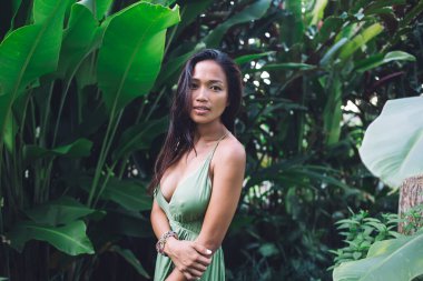 Çekici, uzun saçlı, zeytin tenli, açık tenli yeşil dekolte elbise ve bilezik takan ve tropikal bitkilerin yanında dururken kameraya bakan Asyalı bir kadın. 