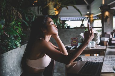 Beyaz bluzlu, ciddi etnik bir bayan. Akıllı telefonuyla selfie çekiyor. Gün ışığında sıcak kafeteryadaki masada oturuyor.