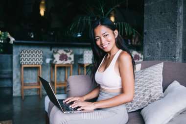 Asyalı pozitif yaratıcı kadın dizlerinin üzerinde dizüstü bilgisayar kullanırken aynı zamanda yazlık kafeteryadaki koltukta kameraya bakıyor.