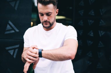 Beyaz gömlekli, kendine güvenen ciddi bir erkek. Bulanık arka planda ofis binasının yakınında beklerken modern akıllı saati kontrol ediyor.