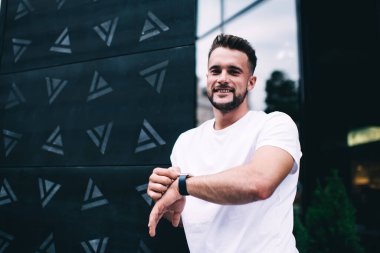 Beyaz tişörtlü mutlu erkek modern akıllı saati kontrol ediyor ve ofis binasının yakınında beklerken kameraya bakıyor.