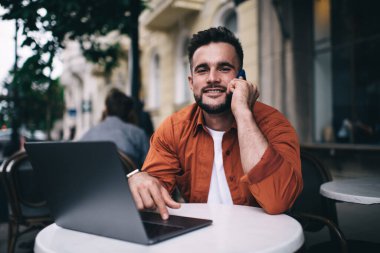 Cafe 'de dizüstü bilgisayarla oturup kameraya gülümserken akıllı telefondan konuşan neşeli genç adam.