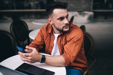 Akıllı telefon kullanarak ve kafeteryada dizüstü bilgisayarla otururken ilgiyle bakışlarını kaçırarak düşünceli genç bir adam.